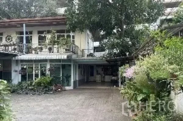Detached House for rent in Watthana, Bangkok - Front courtyard of a two-storey home showing balconies, large windows and shaded parking amid greenery.