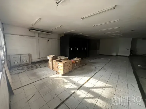 货栈 出租在Khlong Toei, Bangkok - 铺设瓷砖的开放式仓库房间，天花线性灯与堆放的纸箱，旁有配电箱。