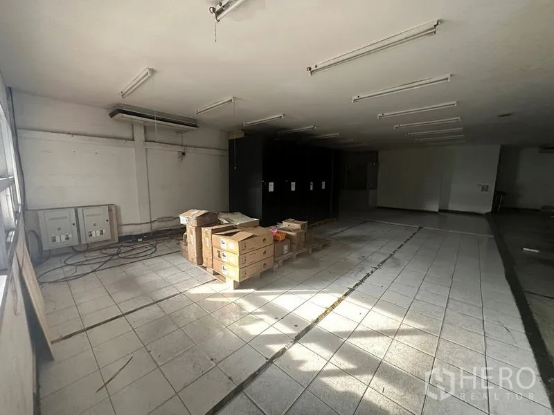 货栈 出租在Khlong Toei, Bangkok - 铺设瓷砖的开放式仓库房间，天花线性灯与堆放的纸箱，旁有配电箱。