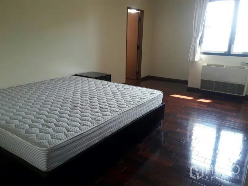 Condominium for rent in Pathum Wan, Bangkok - Large bedroom with big window, wooden floors, bedside table, and wall AC unit.