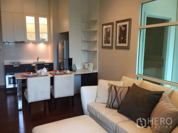 公寓 出租或出售在Watthana, Bangkok - 紧凑型厨房，内置炉灶与白色橱柜，旁边是可供两人用餐的餐桌与舒适沙发。