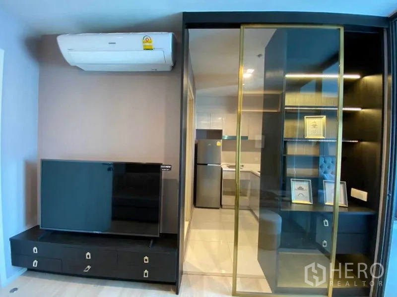 Condominium for rent in Pathum Wan, Bangkok - Living area with TV console and air conditioner, sliding glass to display shelves and kitchen.