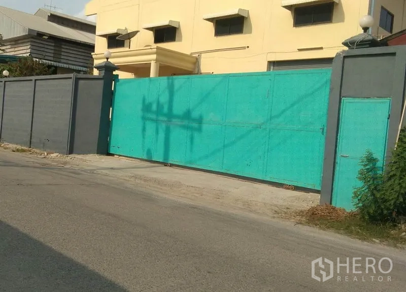 Factory for rent in Bang Phli, Samut Prakan - Secure factory frontage with tall perimeter wall and wide turquoise sliding gate on a quiet Bang Phli road.