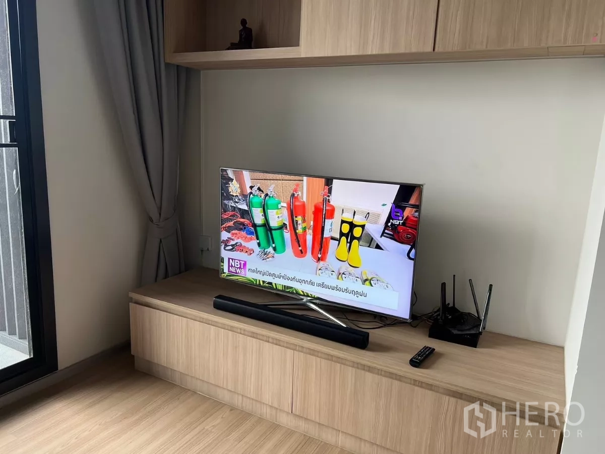 Condominium for rent or sale in Chatuchak, Bangkok - TV on a low media console beside a floor‑to‑ceiling window with grey curtains.