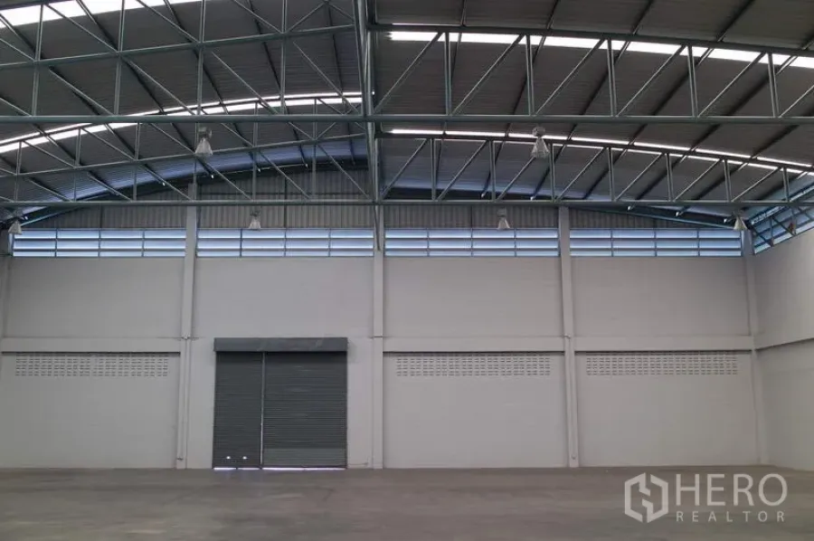 Factory for rent in Pathum Thani - High-ceiling factory warehouse interior with steel truss roof, clerestory vents and a wide roller shutter door.