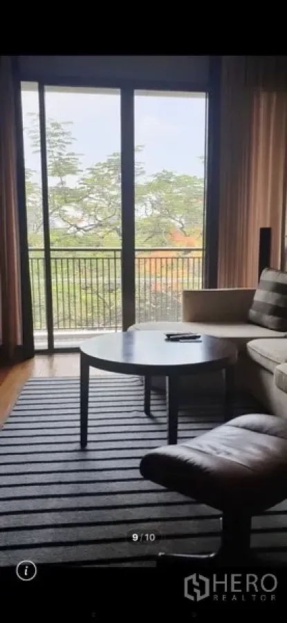 Condominium for rent in Watthana, Bangkok - Cozy living area with sofa, coffee table and striped rug beside floor-to-ceiling sliding doors to a leafy balcony.