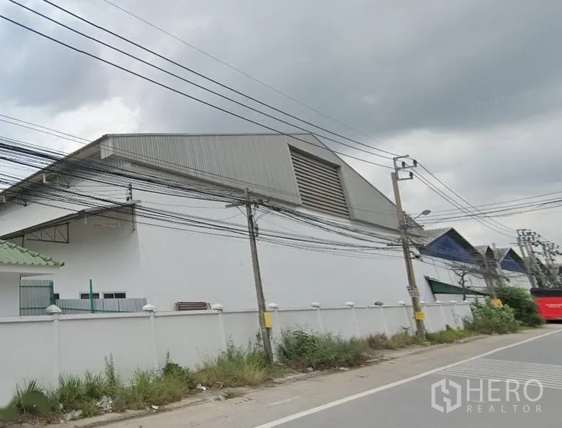 货栈 出租在Samut Prakan - 街景中的大型白色仓库，金属人字形高屋顶带通风百叶，周边有围墙和电线，位于Samut Prakan Bang Sao Thong 工业区。