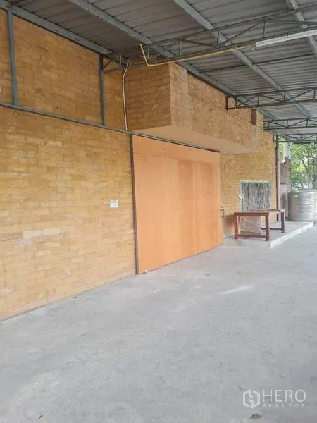 Warehouse for rent in Bang Yai, Nonthaburi - Covered exterior area with brick wall and sliding wooden door beside a concrete floor.