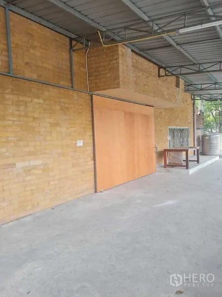 Warehouse for rent in Bang Yai, Nonthaburi - Covered exterior area with brick wall and sliding wooden door beside a concrete floor.