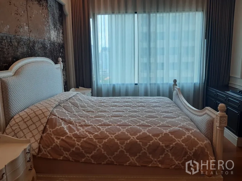 Condominium for rent in Khlong Toei, Bangkok - Cozy bedroom with upholstered bed, patterned bedding, and a large window with skyline outlook.