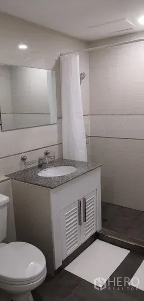 Condominium for rent in Khlong Toei, Bangkok - Light-toned bathroom with granite-top vanity, mirror and shower area with curtain.