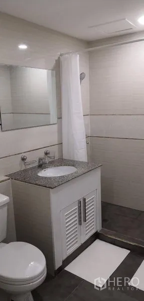 Condominium for rent in Khlong Toei, Bangkok - Light-toned bathroom with granite-top vanity, mirror and shower area with curtain.