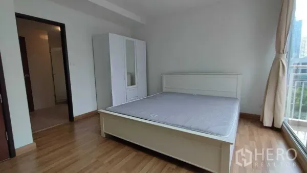 Condominium for rent in Khlong Toei, Bangkok - Sunlit bedroom with white bed frame, wardrobe and full-height window overlooking the Bangkok skyline.