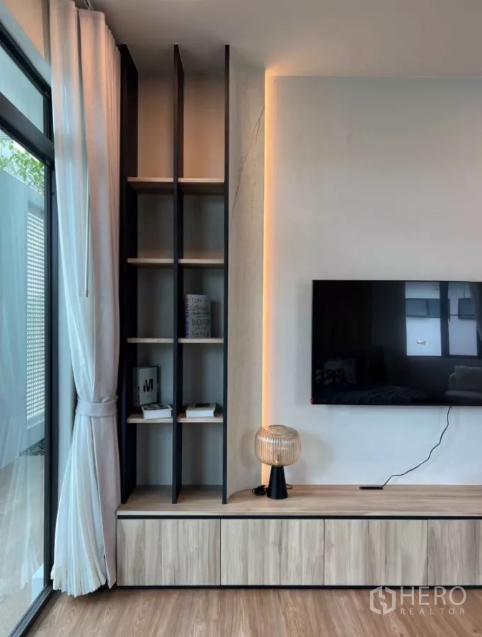 Detached House for rent in Samut Prakan - TV wall with open shelving, warm LED strip lighting, low cabinet and sliding glass door with curtains.