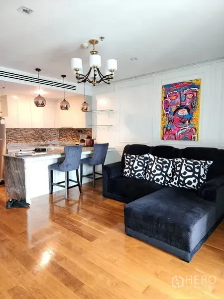 Condominium for rent in Bang Rak, Bangkok - Open‑plan living area with black sectional sofa, marble kitchen island, pendant lights, hardwood floors and colorful wall art.