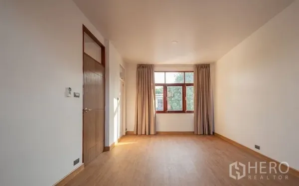 Detached House for sale in Chatuchak, Bangkok - Spacious bedroom with wood flooring, neutral walls and a wide window with curtains.