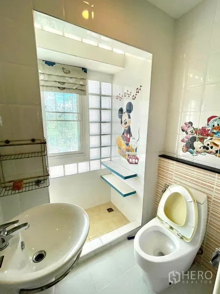 Townhouse for rent in Saphan Sung, Bangkok - Clean bathroom with sink, toilet and glass‑block shower area featuring a playful cartoon tile mural.
