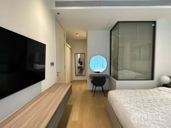 Condominium for rent in Bangkok - Bright Thonglor condo bedroom with wood floors, wall-mounted TV, study desk with round mirror, and glass partition with blinds.