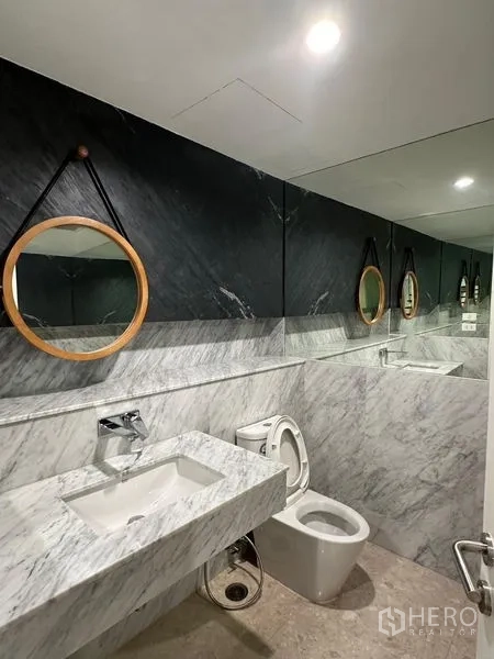 Townhouse for sale in Yan Nawa, Bangkok - Contemporary half-marble bathroom with wall-length mirror, round hanging mirrors, basin and toilet.