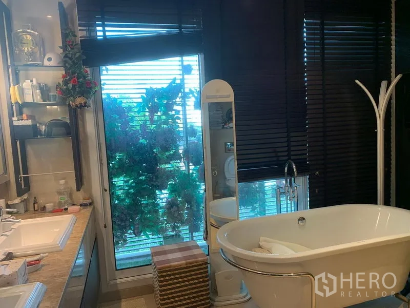 Condominium for sale in Khlong Toei, Bangkok - Bathroom with freestanding soaking tub, double vanity and window blinds with greenery outside.