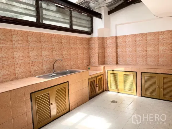 Townhouse for rent in Khlong Toei, Bangkok - Thai-style kitchen with mosaic tiles, stainless-steel sink, louvered cabinets, and high windows.