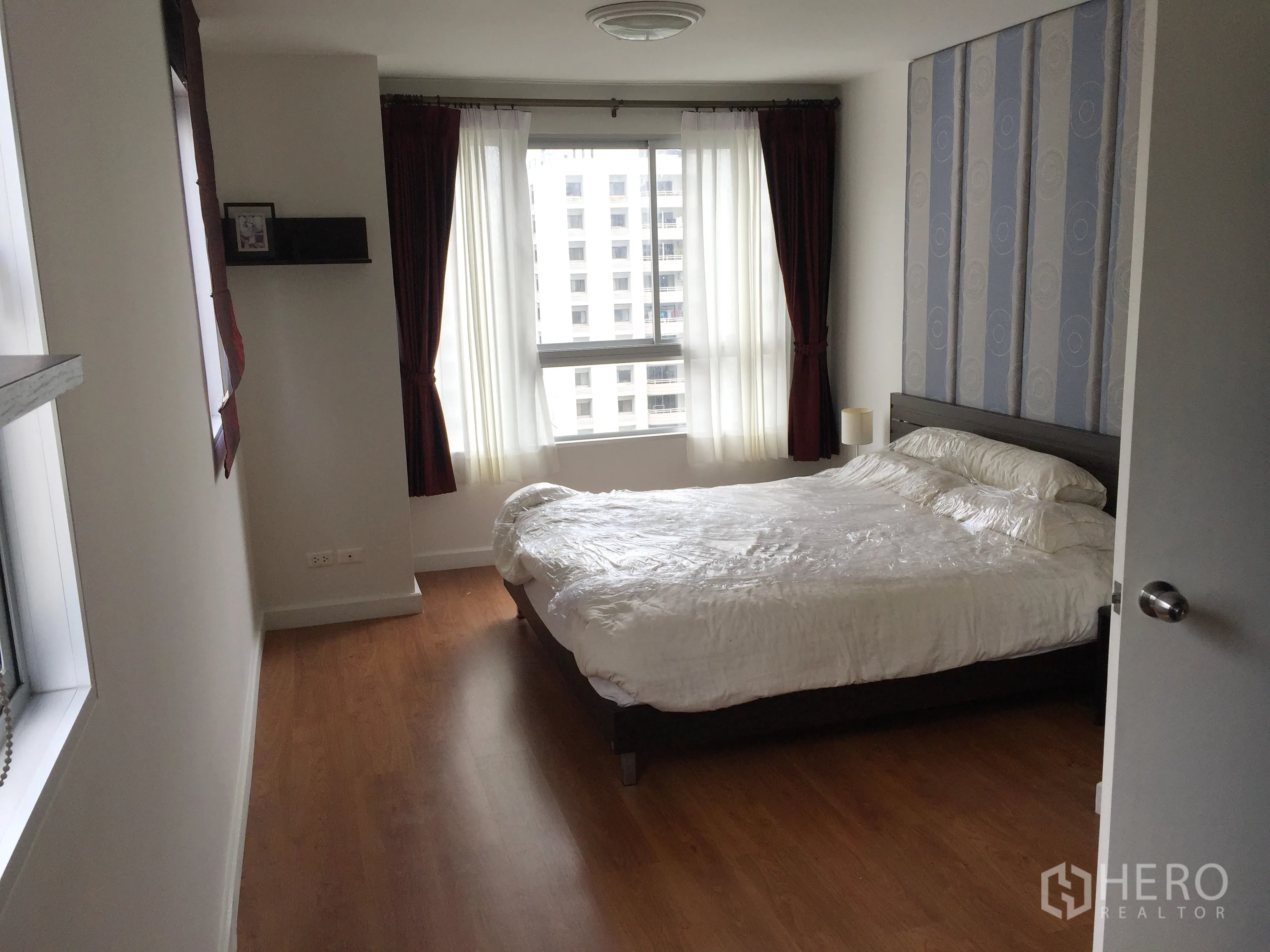 Condominium for rent in Khlong Toei, Bangkok - Bedroom with double bed, large curtained windows, wood flooring and city view.