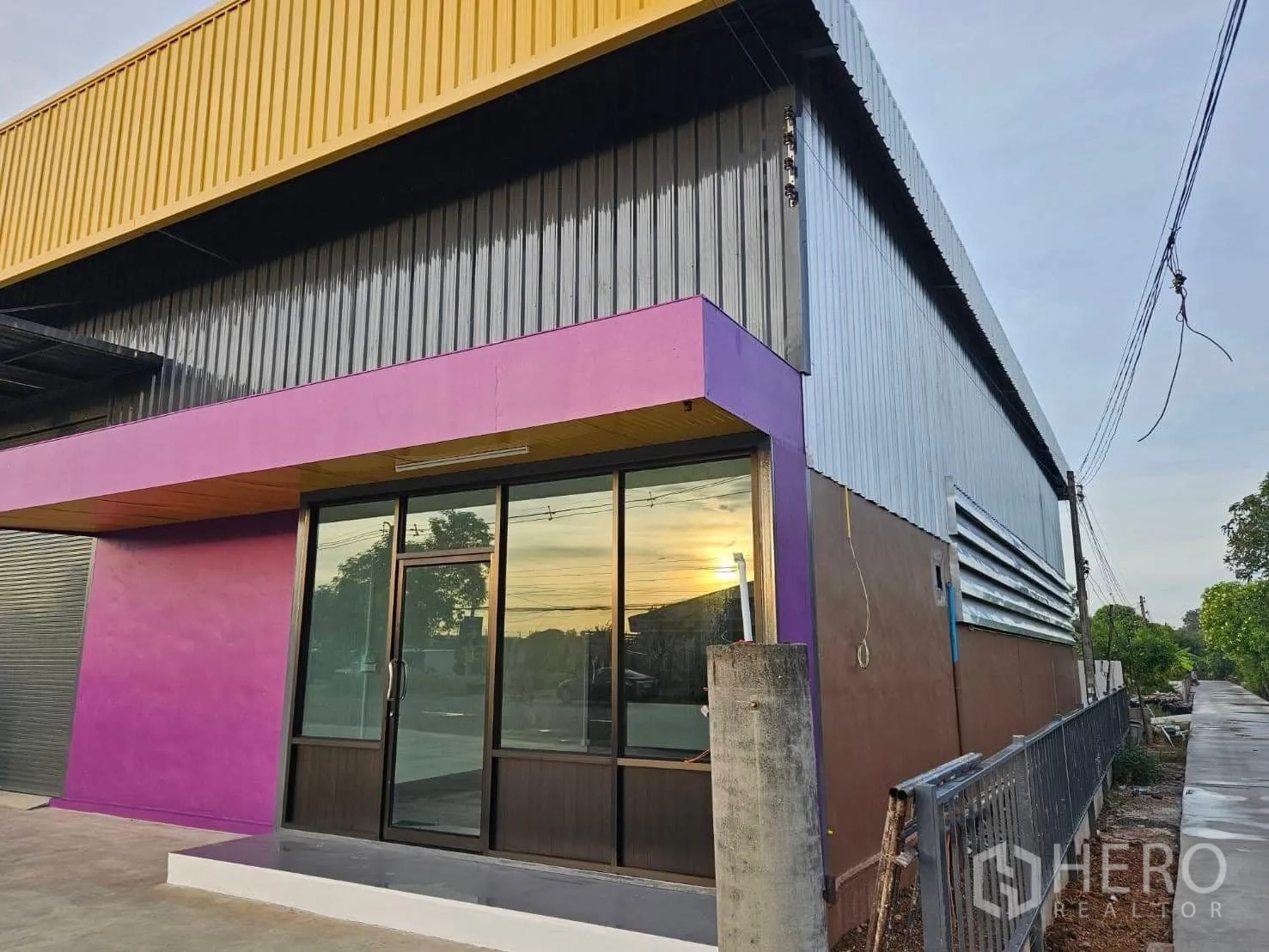 货栈 出租在Lam Luk Ka, Pathum Thani - 建筑转角展示带紫色饰带的玻璃办公室入口与黄色屋檐。