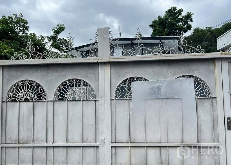 Detached House for rent in Phra Khanong, Bangkok - Decorative white metal gate with arched lattice panels and trees, leading to a detached house under cloudy sky.