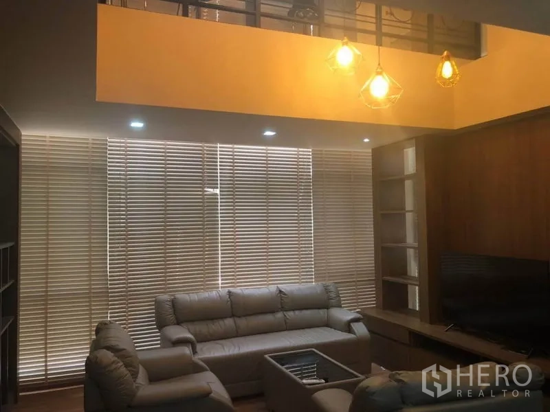 คอนโด ขายในBang Na, Bangkok - ห้องนั่งเล่นเพดานสูง โคมไฟห้อย โซฟาหนัง ตู้วางทีวี และมู่ลี่เต็มผนัง
