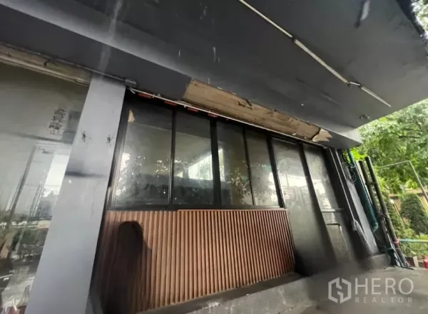 店屋 出租在Khlong Toei, Bangkok - 临街店面配黑色雨棚，大幅玻璃与竖向木饰面，邻近道路绿化。