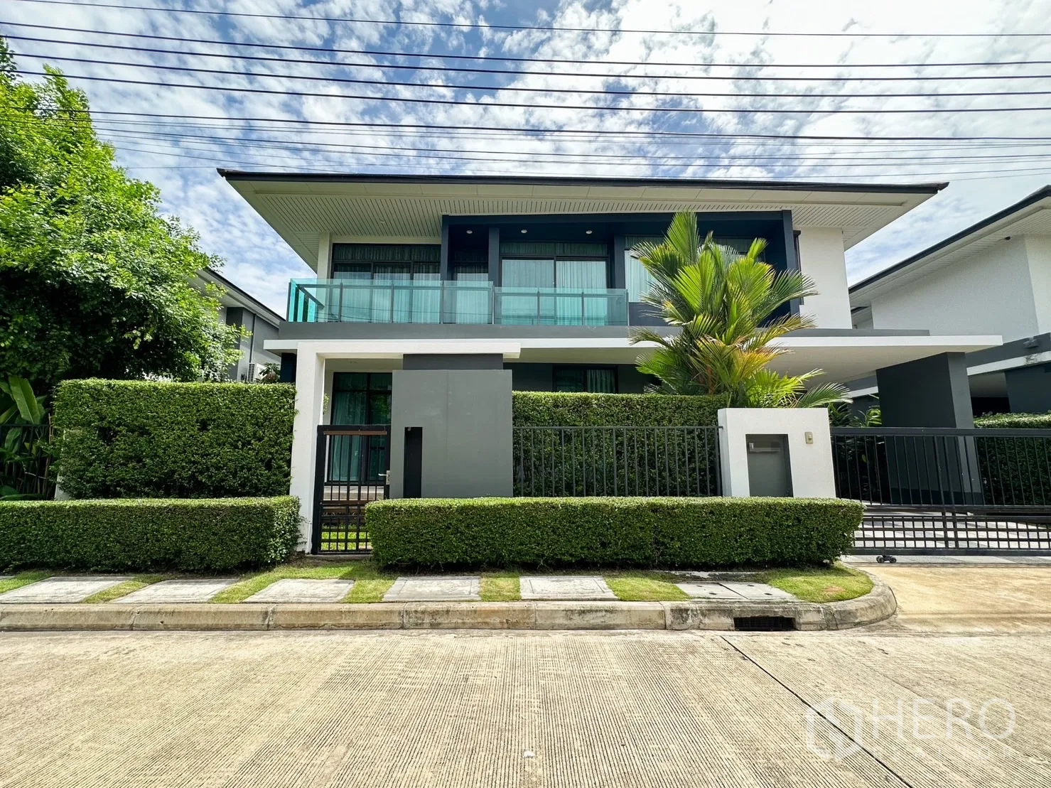 Detached House for rent or sale in Bang Kapi, Bangkok - Modern two-storey detached house facade with glass balcony, manicured hedges and gated frontage under blue sky.