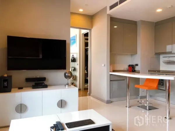 Condominium for rent in Bang Rak, Bangkok - Open-plan living area with wall-mounted TV, glossy kitchen and breakfast bar with an orange stool.