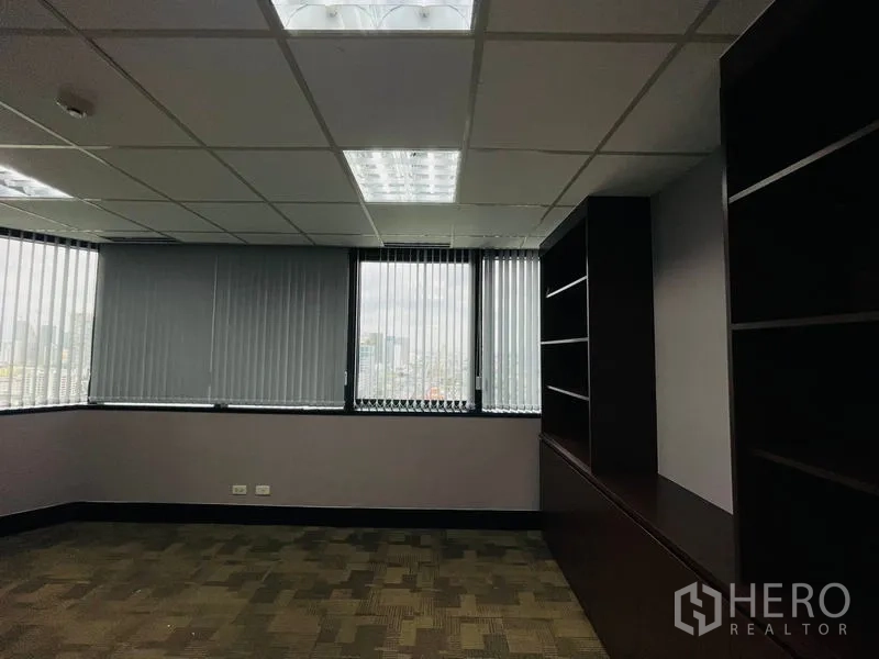 Office Space for rent - Corner executive room with city views, vertical blinds, and dark built‑in shelving under a grid ceiling.