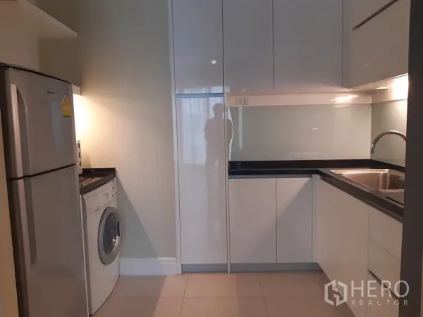Condominium for rent or sale in Khlong Toei, Bangkok - Modern U-shaped kitchen with glossy white cabinets, sink, refrigerator and front-load washing machine.