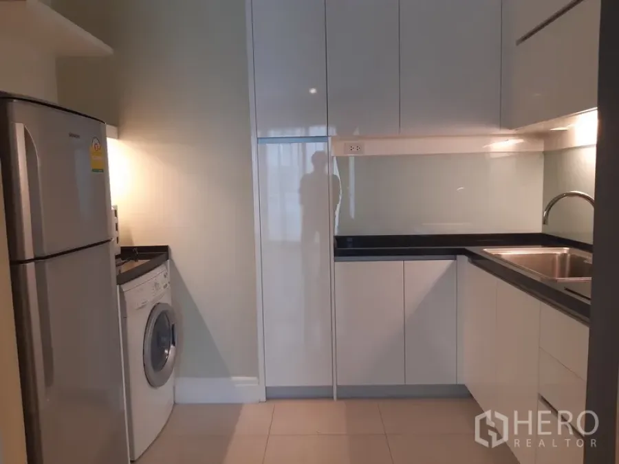Condominium for rent or sale in Khlong Toei, Bangkok - Modern U-shaped kitchen with glossy white cabinets, sink, refrigerator and front-load washing machine.