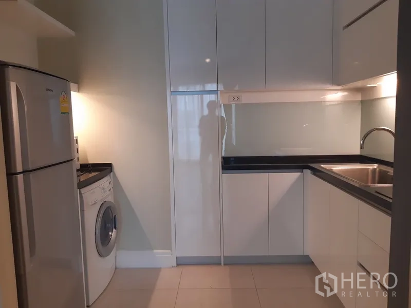 Condominium for rent or sale in Khlong Toei, Bangkok - Modern U-shaped kitchen with glossy white cabinets, sink, refrigerator and front-load washing machine.