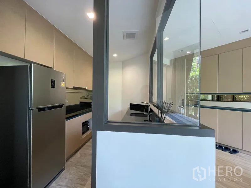 Condominium for sale in Watthana, Bangkok - Separate galley kitchen with glass partition, black countertops, and stainless-steel refrigerator.