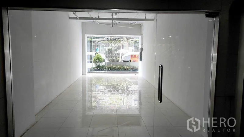 Retail Space for rent in Khlong Toei, Bangkok - Interior view of compact white retail space with polished tiles, track lighting, and front window facing greenery in Khlong Toei, Bangkok.