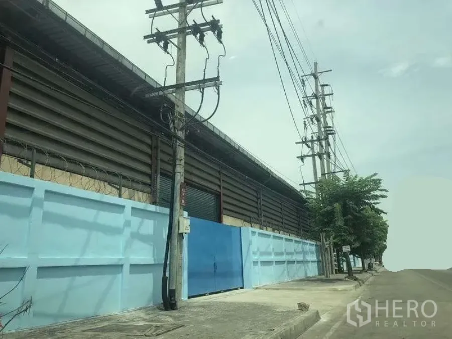 คลังสินค้า ให้เช่าในPhra Pradaeng, Samut Prakan - มุมมองถนนด้านหน้าโกดัง รั้วสีฟ้า ประตูเหล็ก และเสาไฟฟ้าตามแนวถนนกว้าง