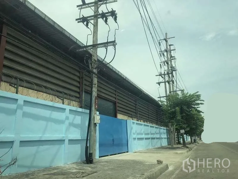 คลังสินค้า ให้เช่าในPhra Pradaeng, Samut Prakan - มุมมองถนนด้านหน้าโกดัง รั้วสีฟ้า ประตูเหล็ก และเสาไฟฟ้าตามแนวถนนกว้าง