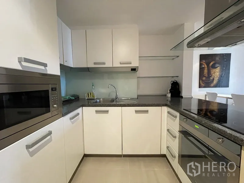 Condominium for rent or sale in Watthana, Bangkok - U-shaped kitchen featuring white cabinetry, stone countertops, oven, cooktop, and sink.