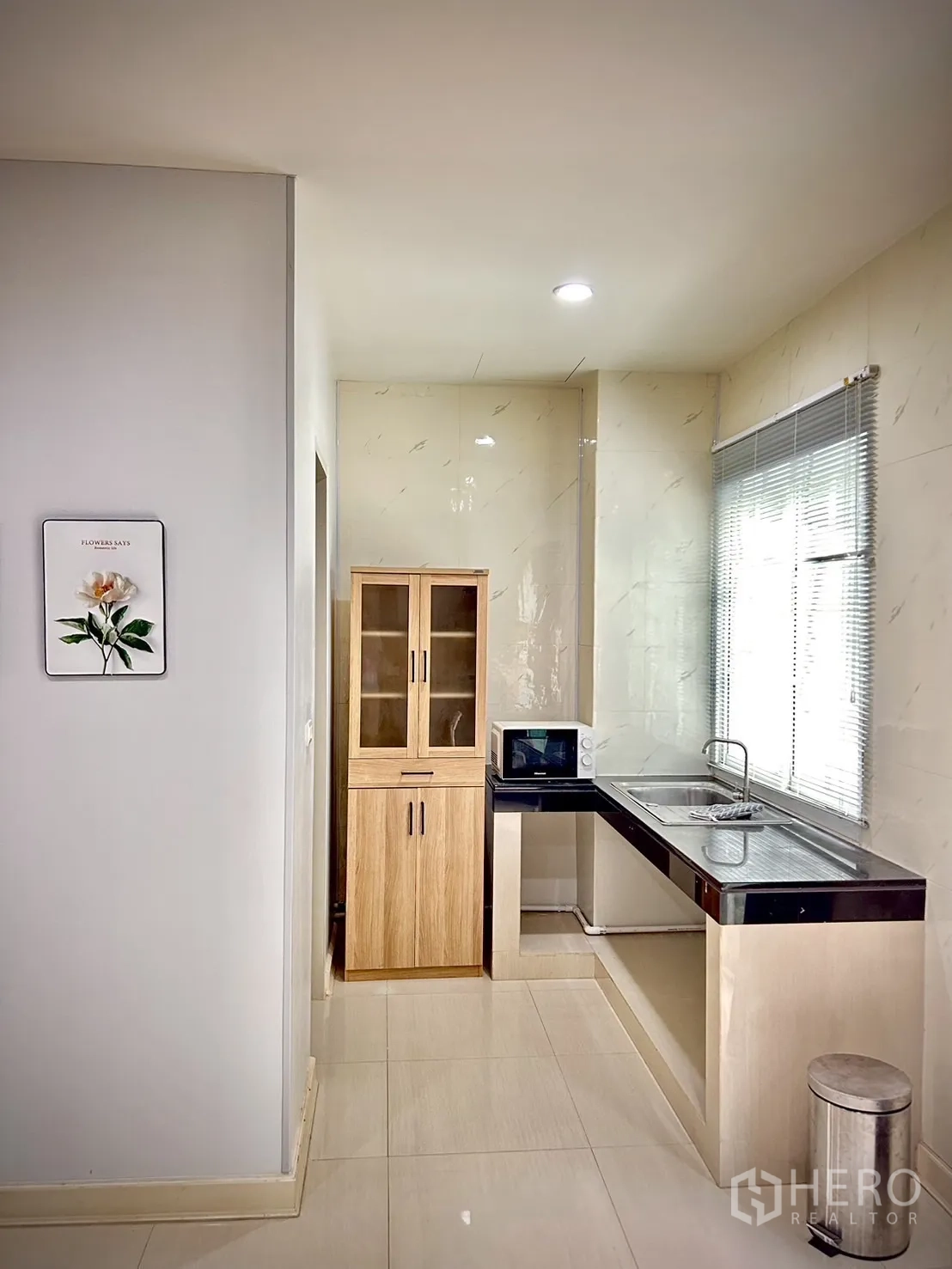 Townhouse for rent in Bang Kapi, Bangkok - Compact light-toned kitchen with window blinds, stainless sink, microwave and wood pantry cabinet.