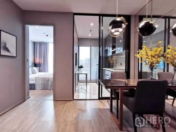 Condominium for sale in Watthana, Bangkok - Open-plan dining area with pendant lights, glass partition to a bright kitchen and balcony, wood floors, and a view toward the bedroom in a Watthana condo near BTS Ekkamai.