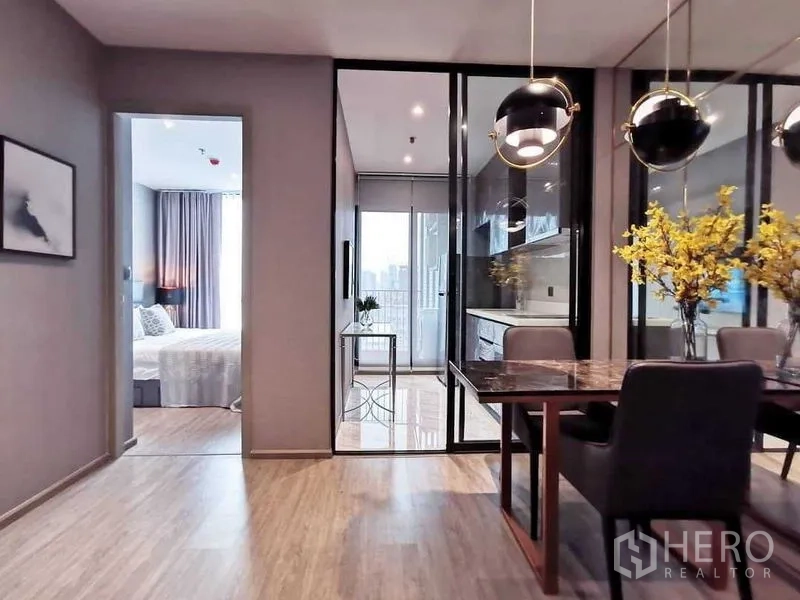 Condominium for sale in Watthana, Bangkok - Open-plan dining area with pendant lights, glass partition to a bright kitchen and balcony, wood floors, and a view toward the bedroom in a Watthana condo near BTS Ekkamai.