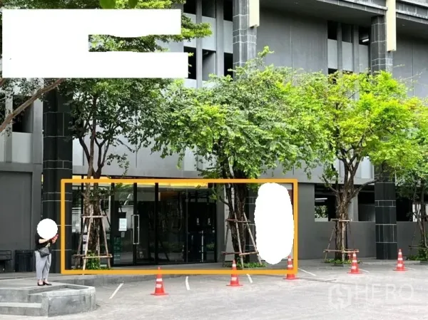 零售空间 出租在Watthana, Bangkok - 现代建筑的一层零售门面，落地玻璃立面，门前有绿树与临时停车位。