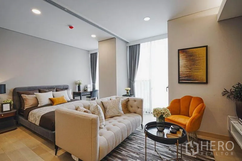 Condominium for sale in Khlong Toei, Bangkok - Bedroom suite with floor-to-ceiling window, beige sofa and an orange accent chair in the living nook.