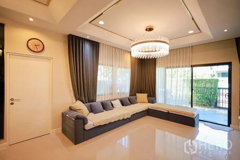 Detached House for rent or sale in Lam Luk Ka, Pathum Thani - Elegant living room with L-shaped sofa, chandelier and floor-to-ceiling windows.