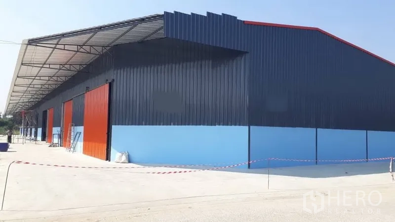 货栈 出租在Pak Kret, Nonthaburi - Pak Kret 现代蓝色钢结构仓库外观，红色滑动门，宽阔混凝土场地与长挑檐装卸区。