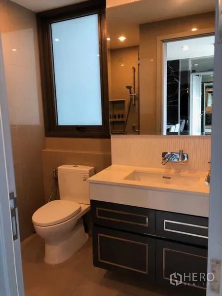 Condominium for rent in Khlong Toei, Bangkok - Modern bathroom with window, vanity, toilet, and glass-enclosed shower.