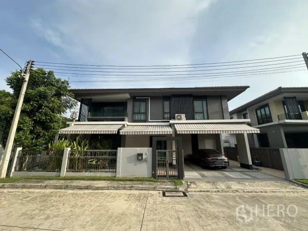 Detached House for rent or sale in Lam Luk Ka, Pathum Thani - Modern two-storey detached house with gated fence, striped awnings and a covered two-car carport on a quiet street in Lam Luk Ka.